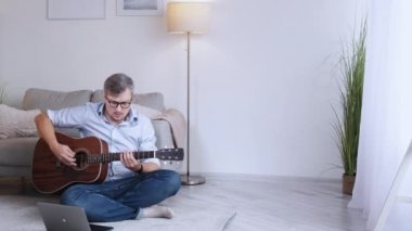Gitar dersi. Erkek oyuncu. Online eğitim. Akustik enstrümanla oynayan zeki, orta yaşlı bir adam yerde dizüstü bilgisayarla oturup fotokopi odası boşluğunda oturuyor..