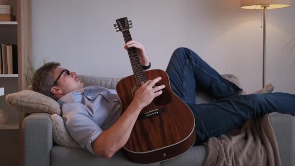 音乐放松 随便的男人 回家休息 漂亮的中年男人在室内放沙发弹奏吉它和作曲曲调 — 图库视频影像