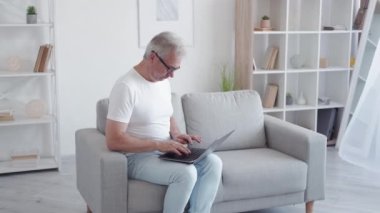 İnternette iş yorgunluğu. Göz yorgunluğu. Bağımsız yaşam tarzı. Yorgun, orta yaşlı bir adam. Gözlüklerini çıkarıyor. Açık modern evdeki koltukta dizüstü bilgisayar kullanıyor..