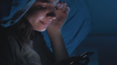 Dijital bağımlılık. Yorgun bir kadın. Çevrimiçi hayat. Uykusuz, gülümseyen kadın yatağında kıvrılıyor akıllı telefon esniyor ışık gölge odasının içinde bitkin hissediyor. Dikey video.