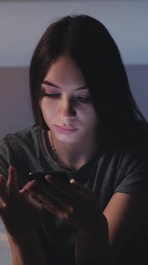 Dijital hayat. Bağımlı kadın. Gezici sohbet. Uykusuz, gülümseyen, yatakta oturan, akıllı telefonları kaydıran, ışık gölge odasının içinde bitkin hisseden kadın. Dikey video.