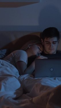 Gece yatağı işi. Bağımsız yaşam tarzı. İnternet uykusuzluğu. Uyuyan, yorgun kocayı kucaklayan bir kadın karanlık odada dizüstü bilgisayar kullanıyor. Dikey video.