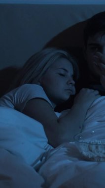 Gece filmi. Uykusuzluk sorunu. Geç saatlere kadar boş vakit. Sıkılmış koca TV izlerken yatakta patlamış mısır yerken yorgun kadın karanlık odada uyuyor. Dikey video.