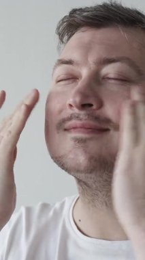 Erkek kozmetikleri. Mutlu adam. Yüz bakımı. Metroseksüel tatmin olan adam, arka planda yüzüne nemlendirici krem sürmekten zevk alıyor. Dikey video.