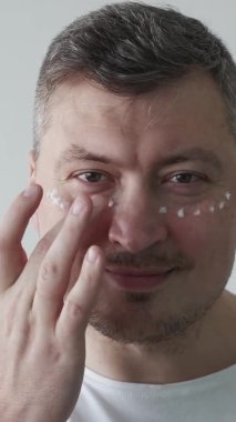 Yaşlanma karşıtı prosedür. Erkek bakımı. Cilt bakımı. Mutlu çekici bir adam, arka planda göz altına nemlendirici krem sürmekten zevk alıyor. Dikey video.