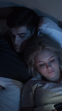 Gadget gecesi. İnternet keyfi. İletişim sorunu. Cep telefonlarını kullanarak yatakta yan yana uzanan yorgun çiftler. Dikey video.