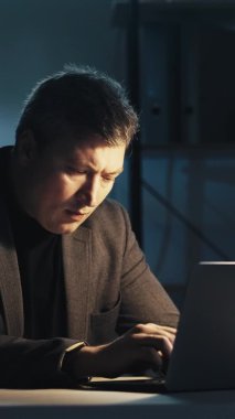 Gece işi. İşkolik yaşam tarzı. Projenin son teslim tarihi. Karanlık ev ofis işyerinde bilgisayarı kullanan dalgın bir iş adamı. Dikey video.
