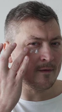 Yüz bakımı prosedürü. Erkek tedavisi. Deri losyonu. Kendine güvenen bir adam, arka planda göz altına nemlendirici krem sürmekten zevk alır. Dikey video.