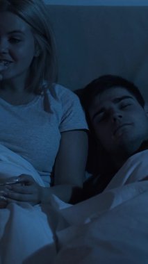 Gece filmi. Geç saatte eğlence. Ev tatili. Heyecanlı, meraklı eş, karanlıkta uyuyan yorgun bir adamla yatakta patlamış mısır yiyen eğlenceli bir TV programı izlemekten zevk alıyor. Dikey video.