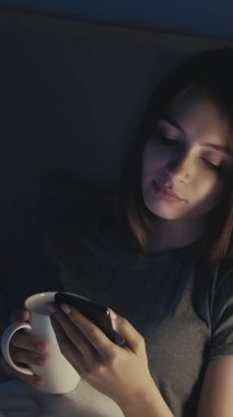 Çevrimiçi bağlantı. Uykusuz kadın. Dikey video. Yatakta güzel bir bayan, akıllı telefonuyla sosyal medyada kayıyor ve aydınlık gölge odasının içinde çay içiyor..