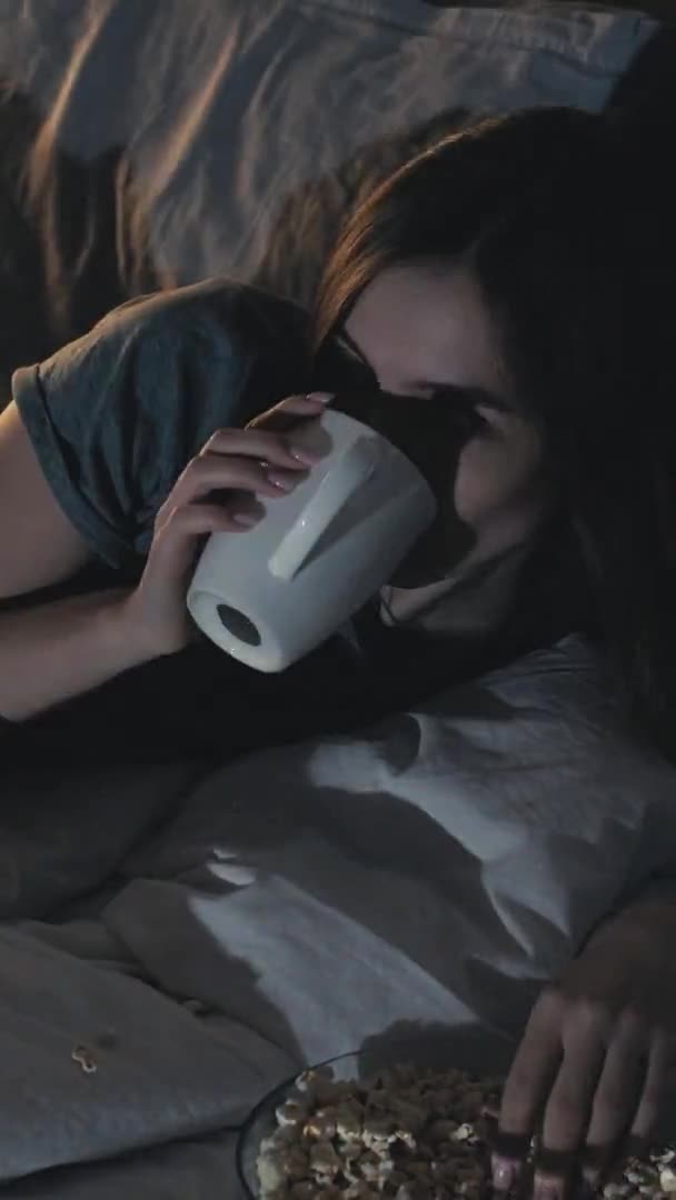 喜剧表演 睡眠不足的夜晚 家庭休闲 快乐的女人看了有趣的电影吃爆米花喝饮料奠定了床黑暗的房间内部 垂直录像 — 图库视频影像