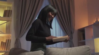 Ev ödevi. Hobi olarak. Edebiyat akşamı. Mutlu, mutlu, zeki bir kadın modern oturma odasının iç kısımlarında sayfaları çevirerek öğrenmenin keyfini çıkarıyor..