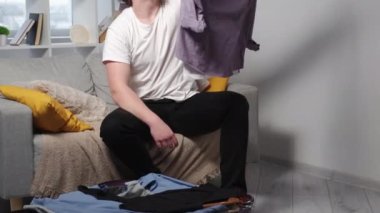 Unpacking suitcase. Happy man. Vacation trip. Smiling relaxed guy sitting sofa taking clothes from baggage after travelling in light home room interior.