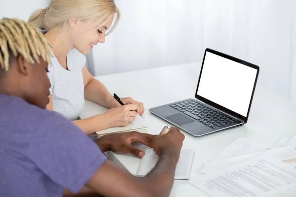 Online education. Studying couple. Digital mockup. Smiling woman and black man making notes in copybooks sitting desk with laptop blank screen in light room interior.