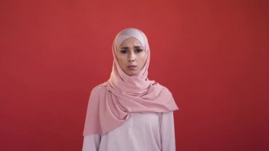 Frightened woman. Omg expression. Shocking news. Shocked speechless worried female face in hijab covering mouth not to scream isolated on red background.