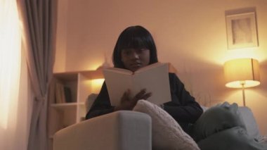 Evde öğrenim. Bilgi bilgeliği. Eğitim hobisi. Zeki düşünceli kadın modern oturma odasındaki koltukta açık kitap okumaktan zevk alıyor..