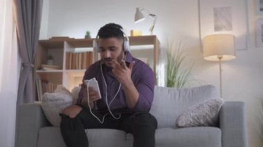 Sanal toplantı. Ev görüntülü konuşma. Çevrimiçi iletişim. Kulaklıklı, akıllı telefon kamerasıyla konuşan, modern oturma odasındaki rahat koltukta oturan kendine güvenen bir adam..