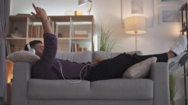 Music energy. Positive vibe. Home leisure. Relaxed amused guy enjoying listening playlist online in headphones using phone lying on couch at modern living room interior.