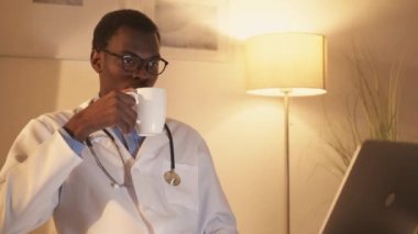 Medical webinar. Healthcare training. Telemedicine consulting. Focused male doctor watching video class on laptop drinking tea late at modern workplace interior.