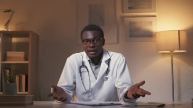 Teletıp görüntülü arama. Sanal randevu. Dijital klinik. Modern iç mekanda geç saatlere kadar çalışan cahil erkek doktor omuz silkiyor..