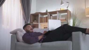 Web chat. Home video meeting. Digital leisure. Relaxed man enjoying conversation using notebook lying on cozy couch at modern living room interior.