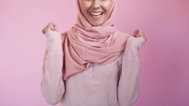 Kutlayan kadın. Başarı sevinci. Gol başarısı. Heyecanlı, mutlu, mutlu, dişlek, tesettürlü, tesettürlü, tesettürlü, pembe arka planda baş parmak gösteren kadın..
