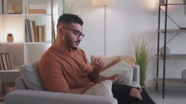 Reading leisure. Home rest. Literature hobby. Smart curious man in glasses sitting with open book turning pages on couch at modern living room interior.