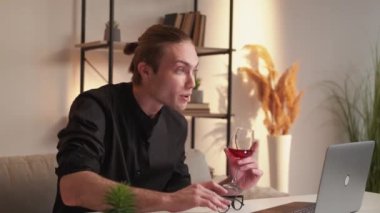 Drinking online. Video call. Virtual toast. Distance communication. Relaxed confident business man with glass of red wine speaking on laptop camera at modern home workplace interior.