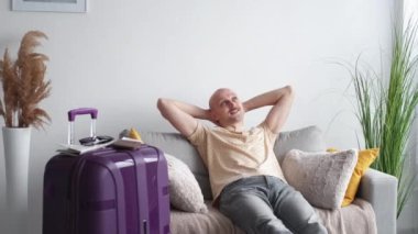 Vacation holiday. Relaxed man. Enjoying moment. Happy dreamful guy resting on sofa with travelling stuff and suitcase in light room interior.
