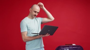 Choose trip. Concentrated man. Online order. Puzzled guy looking laptop in hands getting positive news staying with suitcase on red background.
