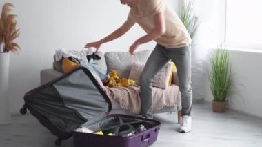 Vacation packages. Playful man. Holiday excitement. Unrecognizable happy funny guy putting clothes stuff into suitcase in light room interior.