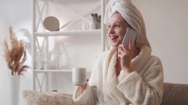 Fun phone talk. Home leisure. Mobile communication. Excited amused laughing woman in bathrobe with coffee mug enjoying conversation at modern living room interior.