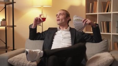 Lüks bir hayat. Zengin insanlar. Zenginlik başarısı. Memnun milyoner adam dolar parasıyla rahatlıyor. Modern iç mekanda kırmızı şarap içiyor..