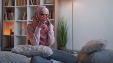 Kahve gevşedi. Samimi bir kızlık zarı. Hafta sonu evde. Keyifli mutlu kadın tesettürlü rahat yastıkların üzerinde bir bardak sıcak içecek içiyor oturma odasında..