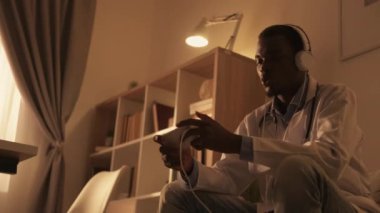 Doctor break. Online entertainment. Digital fun. Excited male medical worker in headphones enjoying playing video game on phone after work on couch in modern interior.