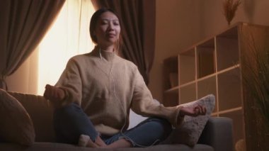 Hafta sonu meditasyonu. Sakinleştirici terapi. Farkındalık zen. Nilüfer çiçeğiyle yoga yapan huzurlu bir kadın Hygge Home 'daki kanepede oturup kulaklıkla müzik dinliyor..