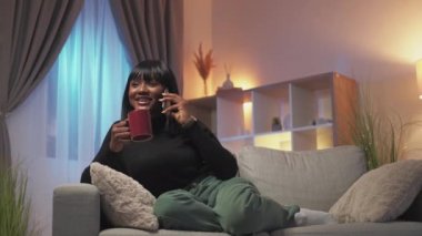 Home phone call. Evening rest. Mobile communication. Relaxed amused smiling woman enjoying conversation drinking tea on couch in modern living room interior with free space.