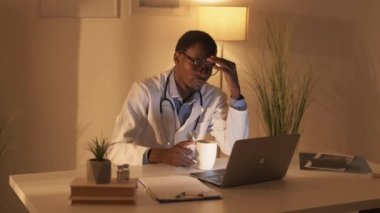 Çok çalışan doktor. Hayat tarzını değiştiriyor. Gece vardiyası yorgunluğu. Yorgun erkek doktor laptopta internetten webinar izleyip koyu renk işyerinde geç saatte kahve içiyor..