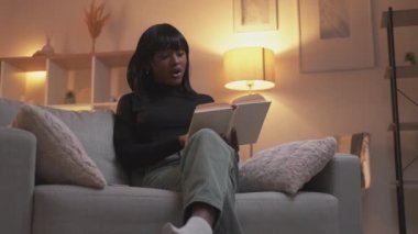 Okuma hobisi. Ev tatili. İyi akşamlar. Rahat, meraklı, akıllı bir kadın, açık bir kitapla modern Hygge oturma odasındaki rahat kanepenin sayfalarını çeviriyor..