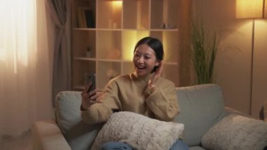 Ev görüntülü arama. Çevrimiçi iletişim. Haftasonu dijital eğlence. Rahat mutlu kız, modern oturma odasındaki rahat koltukta telefonla sanal sohbetten zevk alıyor..