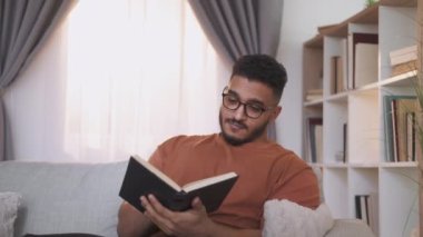 Reading hobby. Book lover. Weekend leisure. Relaxed curious smart guy in glasses enjoying funny story turning pages on home couch at modern interior.