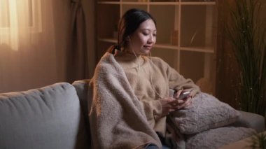 Müzik ilhamı. Kulağa hoş geliyor. Ev tatili. Rahat kız, Hygge 'ın oturma odasındaki rahat koltukta telefon kullanarak kulaklıkla mp3 dinlemenin keyfini çıkarıyor..