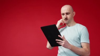 Choose product. Puzzled man. Mobile technology. Curious doubtful guy typing tablet computer making decision to get order red background copy space.