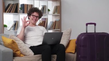 Online tour. Satisfied man. Vacation inspiration. Happy guy with laptop on knees showing ok gesture sitting sofa with packed suitcase in light room interior.
