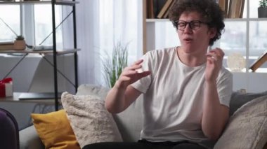 Winner excitement. Expressive man. Vacation joy. Happy guy in spectacles sitting sofa with packed suitcase showing winner fun emotion in light room interior.