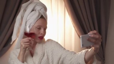 Toast selfie. Virtual celebration. Luxury life. Social media. Happy smiling blogger woman in bathrobe posing with red wine glass on phone camera at home interior.