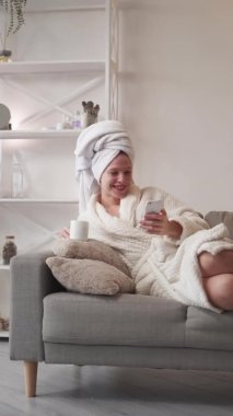 Vertical video. Mobile chat. Gadget weekend. Online home leisure. Relaxed amused laughing woman in bathrobe with coffee mug using phone on couch at modern living room interior.