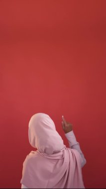 Pointing woman. Promotion advice. Content advertising. Back view of reading female in hijab demonstrating with finger red empty space recommendation background vertical.