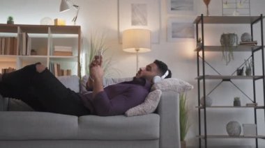 Ev görüntülü arama. Mesafe iletişimi. Hafta sonu tatili. Kulaklıklı rahat adam telefonda konuşuyor. Modern oturma odasının iç tarafındaki rahat koltukta uzanıyor..