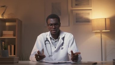 Tıbbi video danışmanlığı. Mesafe tanılama. Klinik tele-sağlık. Kafası karışmış erkek doktor omuz silkiyor. Hasta teşhisi üzerinde çalışıyor. Akşamları modern ev dekorasyonu..